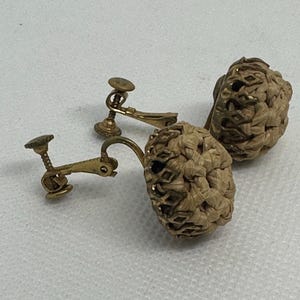 May include: Vintage gold-tone clip-on earrings with woven, beige-colored orb-shaped drops. The earrings have a detailed, textured design and a classic, elegant style. The clip-on mechanism is visible.