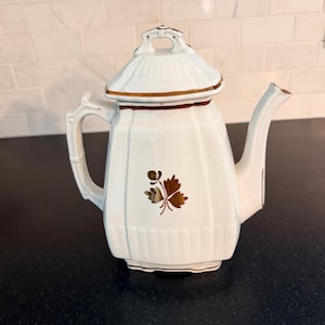 May include: White ceramic teapot with a lid and a handle. The teapot has a brown leaf design and brown trim. The teapot is on a black surface.