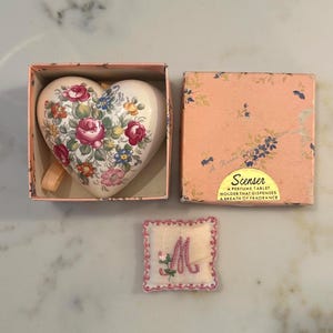 May include: A heart-shaped perfume tablet in a box, featuring a floral design with pink and red roses, blue and yellow accents. The box is peach-colored with floral patterns. A small embroidered pillow with the letter "M" is also present.