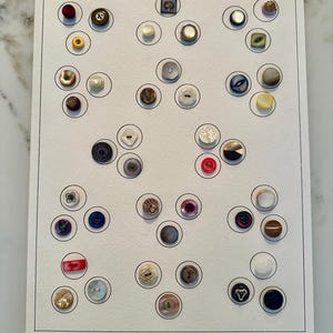 Könnte beinhalten: Eine Sammlung verschiedener Knöpfe, angeordnet auf einer weißen Karte. Die Knöpfe sind in verschiedenen Farben, Formen und Größen gehalten, darunter Rot, Gold, Schwarz und Perlmutt. Die Karte ist mit "Shell" in der oberen linken Ecke beschriftet.