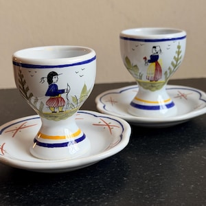 Könnte beinhalten: Zwei weiße Keramik-Eierbecher mit passenden Untertassen. Jeder Becher hat ein handgemaltes Design einer Person in traditioneller Kleidung mit blauen, roten und gelben Akzenten. Die Untertassen haben einen gewellten Rand und ein Kreuzmuster.