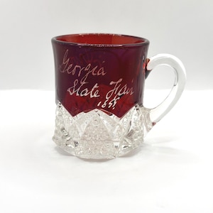 May include: A ruby red glass mug with a clear handle and a cut-glass base. The mug features the inscription "Georgia State Fair 1855" in white script. The mug is likely a vintage collectible.