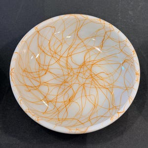 May include: A round, white glass bowl featuring a delicate, intricate pattern of thin, swirling orange lines that crisscross across the interior surface, creating a web-like design that extends from the center to the smooth, rounded rim.
