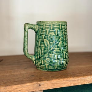 Pode incluir: Uma caneca de cerâmica verde com uma alça. A caneca tem um design de cesta tecida texturizada com detalhes de folhas e flores. A caneca está em uma superfície de madeira.