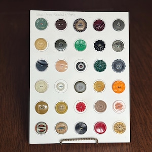 May include: A collection of various buttons in different colors, shapes, and sizes, arranged on a white card. The buttons include shades of green, brown, black, red, and yellow. The card has the text "Sew Thrus- Celeloid & Plastic".