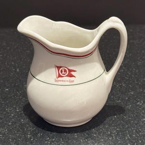 May include: White ceramic creamer with a curved handle and spout. The creamer has a red and green trim, and a red flag emblem with a white "L" and the text "LUCKENBACH LINE".