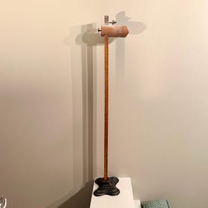 Puede incluir: Un soporte antiguo de madera y metal con una base marrón oscuro. El soporte tiene un poste de madera largo y delgado y una parte superior cilíndrica de color marrón claro con herrajes metálicos. El soporte probablemente se utiliza para exhibición o soporte.