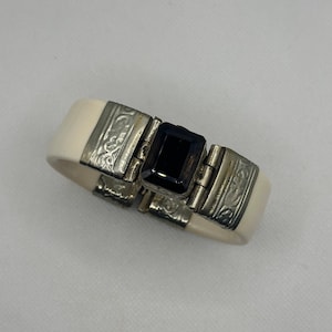 May include: Ivory-coloured hinged bracelet with silver-tone metal accents. The bracelet features a large, rectangular, dark gemstone at its centre. The metal sections have an etched design. A vintage-style accessory.
