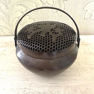 Puede incluir: Un quemador de incienso vintage de metal oscuro con tapa perforada y asa. La tapa presenta cortes florales decorativos. El quemador tiene un cuerpo redondeado y se asienta sobre una superficie blanca desgastada. El asa es arqueada.