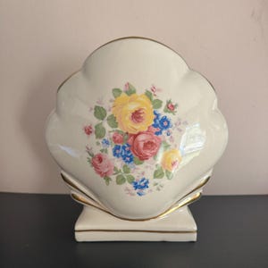 May include: A cream-colored, shell-shaped vase with a floral design. The flowers are yellow, pink, and blue, with green leaves. The vase has gold trim and a rectangular base. The vase is likely ceramic.