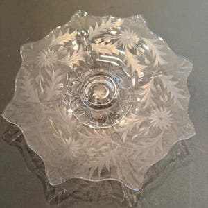 May include: A clear glass serving plate featuring an etched floral pattern with delicate leaves and star-shaped blossoms. The plate has a scalloped, wavy edge and a circular central base with a starburst design etched into the center of the glass.
