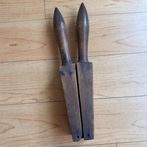 Könnte beinhalten: Zwei antike Holzwerkzeuge mit spitz zulaufenden Griffen und rechteckigen Klingen. Die Werkzeuge bestehen aus dunkelbraunem Holz und weisen Gebrauchsspuren auf. Sie liegen nebeneinander auf einer hellbraunen Holzoberfläche.