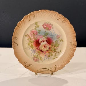 Puede incluir: Un plato de porcelana vintage con borde festoneado y diseño floral. El plato presenta un fondo color crema con un ramo central de rosas rosas y blancas, acentuado por follaje azul y verde, y ribete dorado.