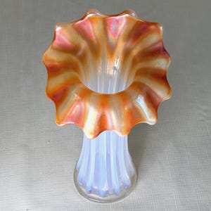 May include: An iridescent glass vase with a ruffled rim. The rim is orange and pink, while the body is pale blue with vertical ridges. The vase has a flared, scalloped top and a rounded base.