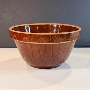 Pode incluir: Uma tigela de cerâmica marrom com um padrão decorativo. A tigela tem uma borda larga e uma forma arredondada. O exterior apresenta uma série de linhas curvas verticais. A tigela é provavelmente usada para preparação de alimentos.