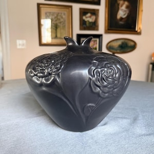 Puede incluir: Un jarrón de cerámica gris oscuro con forma de granada y diseños de rosas en relieve. El jarrón tiene un acabado brillante y dos pequeñas protuberancias en la parte superior. Los detalles florales son prominentes.