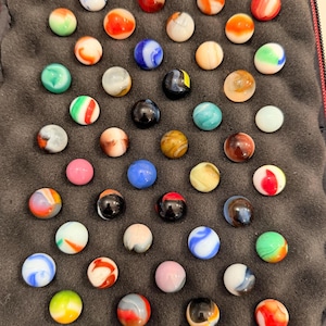 Puede incluir: Una colección de cuarenta y ocho canicas de vidrio variadas dispuestas en un patrón de cuadrícula dentro de un estuche de transporte forrado con espuma negra. Las canicas presentan varios colores, incluyendo rojo, azul, verde, naranja, blanco, negro y marrón.