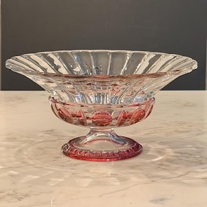 May include: A clear glass bowl with a flared, scalloped rim and a pedestal base. The bowl features a pink accent around the base and rim. The bowl is likely used for serving or display purposes.