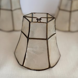 May include: A set of small, off-white lamp shades with a geometric design. Each shade features a light-colored, translucent material encased in a dark metal frame. The shades have a flared, hexagonal shape.