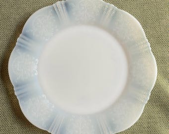 May include: A white, scalloped-edge plate with a floral design around the rim. The plate has a slightly iridescent quality, with a pale blue hue along the edges. The plate is likely made of glass or ceramic.