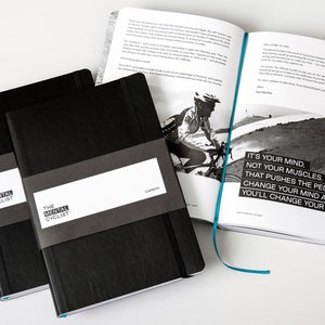 May include: Two black notebooks with the text "The Mental Cyclist" on the cover. The notebooks are closed and stacked on top of each other. The top notebook has the word "CAMBON" printed on the cover. The notebooks are sitting on a white surface. An open book with a black and white photo of a cyclist is in the background.
