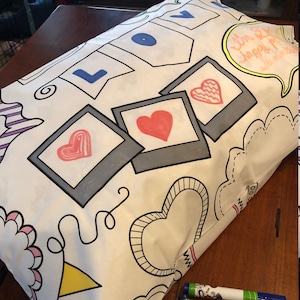 May include: A white pillowcase with a black outline of hearts, swirls, and a banner that says "LOVE". The pillowcase also has a speech bubble that says "I'm so happy you're here!" and several blank picture frames with hearts inside. The pillowcase is lying on a wooden surface with several colorful markers.