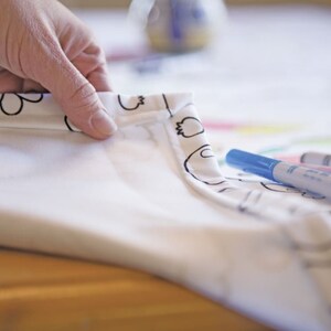 May include: A white tablecloth with a black and white pattern of flowers and other designs. The tablecloth is folded over and a hand is holding the corner. There are three blue, green, and pink markers on the tablecloth.