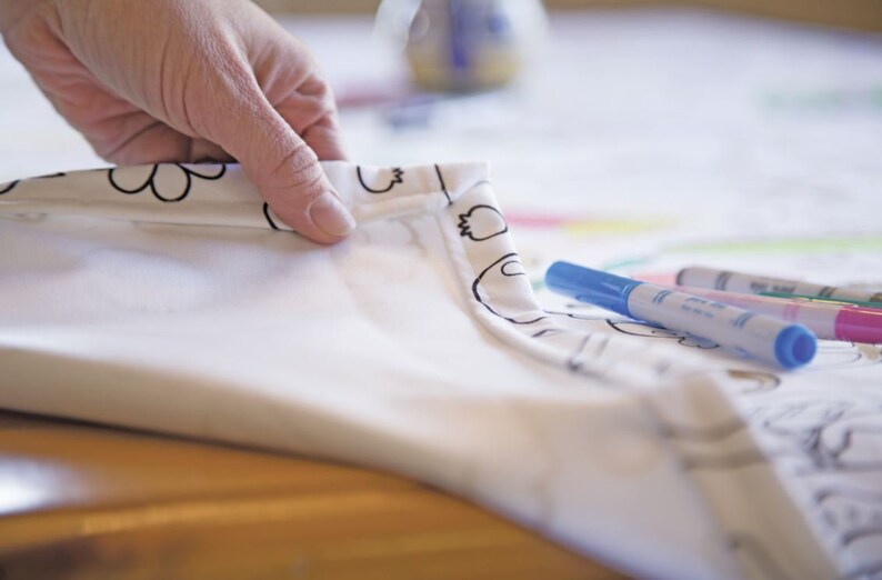 May include: A white tablecloth with a black and white pattern of flowers and other designs. The tablecloth is folded over and a hand is holding the corner. There are three blue, green, and pink markers on the tablecloth.