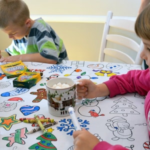 May include: Two children color a white tablecloth with Christmas-themed illustrations. The tablecloth has images of snowmen, Christmas trees, gingerbread men, and other holiday symbols. The children are using markers and crayons to color the images. There is a mug of hot chocolate with marshmallows on the table.