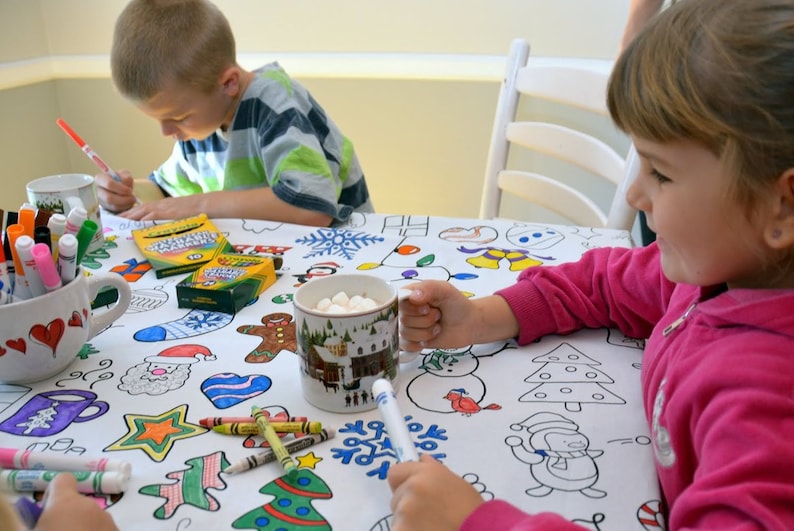 May include: Two children color a white tablecloth with Christmas-themed illustrations. The tablecloth has images of snowmen, Christmas trees, gingerbread men, and other holiday symbols. The children are using markers and crayons to color the images. There is a mug of hot chocolate with marshmallows on the table.