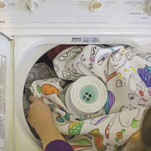May include: A white washing machine with a colorful, hand-drawn design on the fabric inside. The fabric has images of carrots, grapes, and other fruits and vegetables.