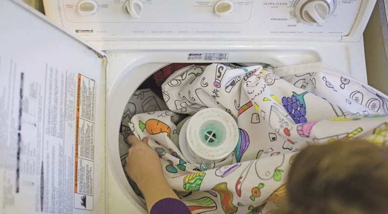 May include: A white washing machine with a colorful, hand-drawn design on the fabric inside. The fabric has images of carrots, grapes, and other fruits and vegetables.