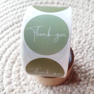 May include: A roll of circular stickers featuring a sage green background and the words "Thank you" in white script. The stickers are on a white backing and are displayed on a small wooden stand.