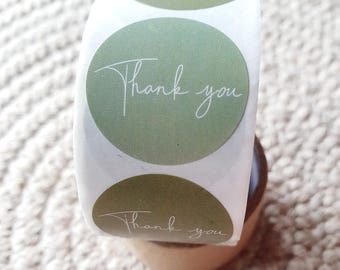 May include: A roll of round stickers with a sage green background and the words "Thank you" in a delicate white script. The stickers are on a white backing and are displayed on a small wooden stand.