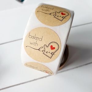 May include: A roll of circular kraft paper stickers. Each sticker displays the words "baked with love" in a handwritten font, alongside a graphic of a mitten with a red heart. These stickers are designed for baked goods.