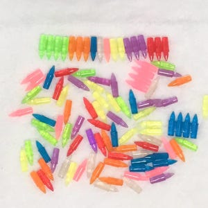 May include: A collection of colorful, translucent plastic toy bullets in various shades of green, orange, blue, yellow, pink, purple, and red. The bullets are scattered across a white surface, with some arranged in neat rows.