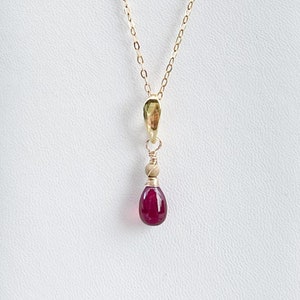 May include: A delicate gold chain necklace with a small, teardrop-shaped pendant featuring a deep red gemstone.