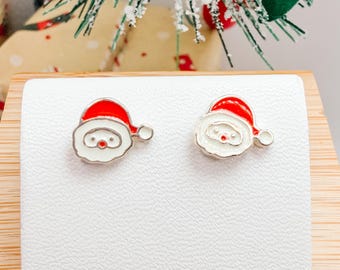 Puede incluir: Un par de pendientes de botón de Papá Noel. Cada pendiente presenta un gorro rojo, una cara blanca y un contorno plateado. Los pendientes se muestran sobre una superficie blanca, con decoraciones navideñas en el fondo.