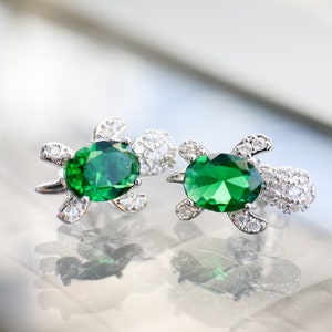May include: A pair of silver turtle earrings with green gemstones. The earrings are set with small clear stones around the edges of the turtle.