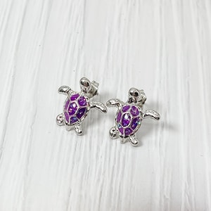 May include: A pair of silver stud earrings featuring a purple turtle design. The turtles have a detailed shell pattern and are a popular choice for jewelry.