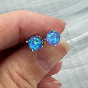May include: A pair of round, blue opal stud earrings. Each earring features a vibrant, iridescent blue stone with hints of green and is set in a silver-coloured metal. The earrings are held in a hand against a light grey background.