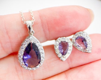 Könnte beinhalten: Eine silberne Halskette mit einem birnenförmigen Anhänger, der einen violetten Edelstein enthält, der von kleineren, klaren Edelsteinen umgeben ist. Passende Ohrringe im gleichen Design sind ebenfalls enthalten.