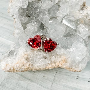 May include: A pair of heart-shaped earrings with vibrant red gemstones set in a silver metal. The earrings are displayed on a white and gray textured surface, showcasing their sparkling facets. A romantic jewelry piece.