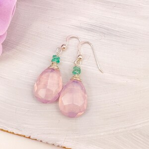 May include: A pair of dangle earrings featuring faceted, light pink teardrop-shaped stones. Each earring has a small, green bead and silver-colored wire and hooks. The earrings are displayed on a white surface.