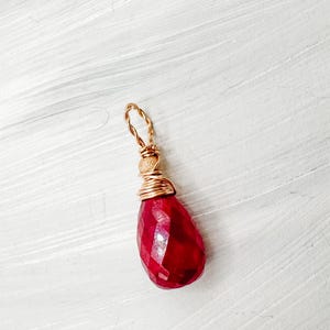 May include: A faceted, teardrop-shaped ruby pendant. The deep red gemstone is wrapped in gold-coloured wire, with a looped top for hanging. The pendant is set against a white background.