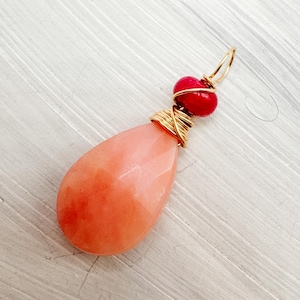 May include: A gemstone pendant featuring a faceted, teardrop-shaped peach-colored stone. Two small red beads and a gold-colored wire wrap the top. The pendant has a gold-colored loop for hanging.