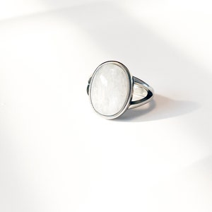 May include: A silver ring featuring a large, smooth, oval-shaped white gemstone set in a polished metal bezel. The band of the ring has a split design that tapers towards the back, showcasing a clean, metallic finish and a reflective, milky stone.