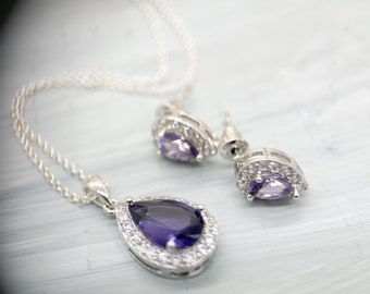 Könnte beinhalten: Ein silbernes Halsketten- und Ohrring-Set mit einem tropfenförmigen violetten Edelstein, der von kleineren klaren Steinen umgeben ist.