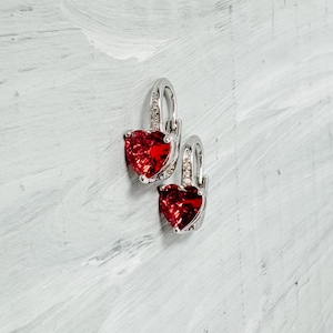 May include: A pair of silver hoop earrings with red heart-shaped gemstones. The earrings have a simple design and are perfect for everyday wear.