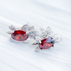 May include: A pair of silver earrings with red gemstone accents. The earrings are shaped like turtles with small, clear stones around the edges.